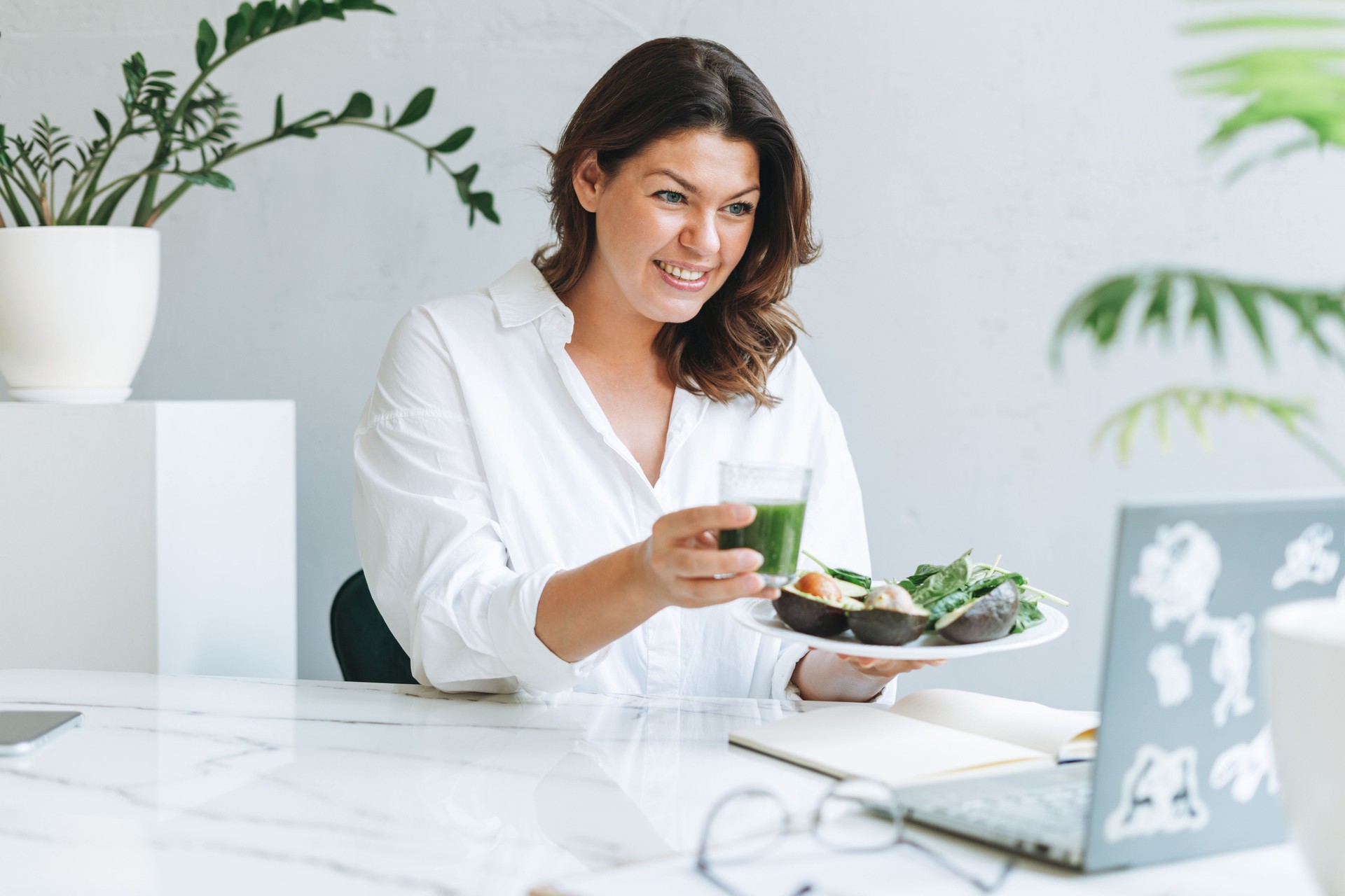 Young smiling brunette woman nutritionist doctor plus size in white shirt with green healthy food at modern bright office room. Doctor communicates with patient online